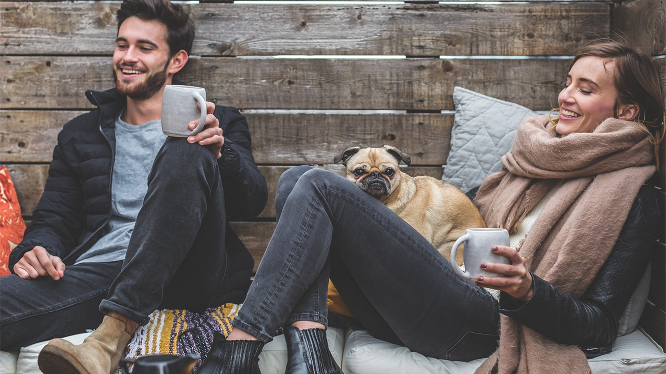 Casal sorrindo com cachorro pug no colo tomando café